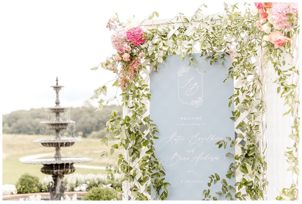 Welcome sign hangs outside of wedding venue with fountain in background. 