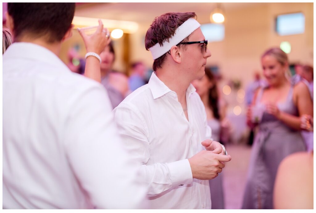 Groomsman with headband looks out into dancing crowd at Equestria West. 